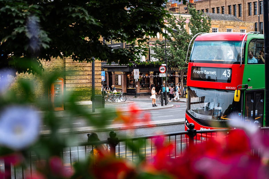 A view of a busy street in Barnet, London, captured during daytime. In the foreground, vibrant pink, red, and white flowers partially obscure the scene, adding a touch of colour. Behind the flowers, there is a black metal railing along the pavement. To the right, a double-decker bus with a green and red exterior is stopped on the road, displaying the route number 43 and destination 'Friern Barnet' on an electronic sign. The street scene features pedestrians walking along the sidewalk, some near bicycles parked on the curb, with others crossing at a pedestrian crossing. The background shows a mix of brick buildings, including shops and pubs, with signage visible, and leafy trees lining the street. The lighting indicates a clear day, typical of urban surroundings in Barnet, as part of a home relocation or furniture transport process, with Man with Van Barnet likely involved in the moving logistics.