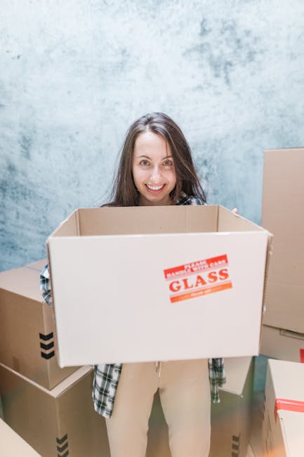 A view of a busy street in Barnet, London, captured during daytime. In the foreground, vibrant pink, red, and white flowers partially obscure the scene, adding a touch of colour. Behind the flowers, there is a black metal railing along the pavement. To the right, a double-decker bus with a green and red exterior is stopped on the road, displaying the route number 43 and destination 'Friern Barnet' on an electronic sign. The street scene features pedestrians walking along the sidewalk, some near bicycles parked on the curb, with others crossing at a pedestrian crossing. The background shows a mix of brick buildings, including shops and pubs, with signage visible, and leafy trees lining the street. The lighting indicates a clear day, typical of urban surroundings in Barnet, as part of a home relocation or furniture transport process, with Man with Van Barnet likely involved in the moving logistics.