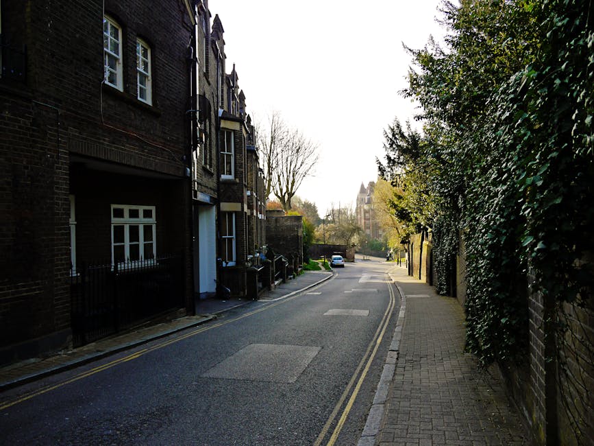 A narrow street scene in High Barnet during daytime, with paved pavement on both sides and a single vehicle lane in the center. On the left, there are small shops with signage, including a barbershop 