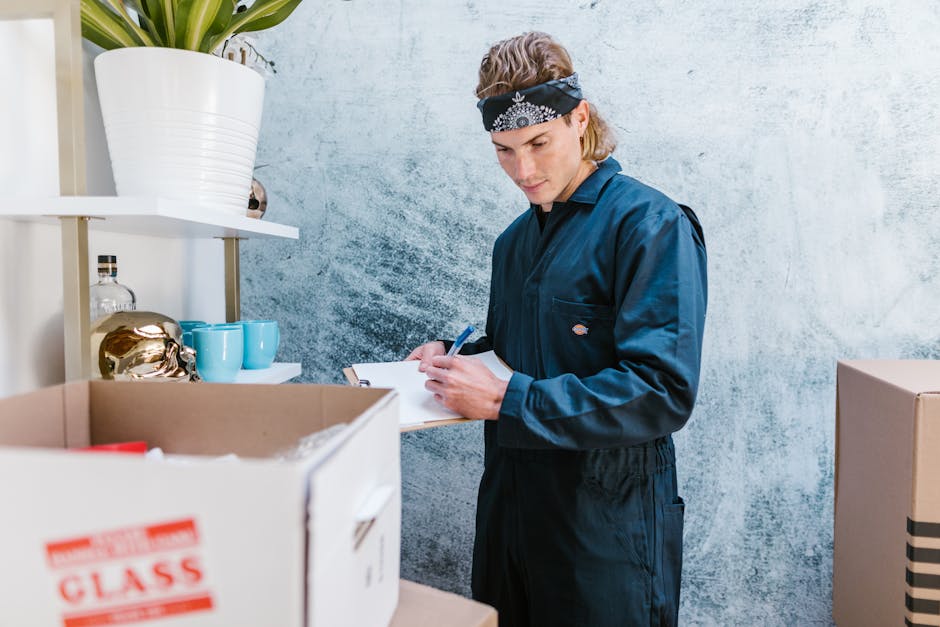 A man dressed in a navy-blue uniform with a black and white patterned headband is inside a room with a textured light blue wall, surrounded by cardboard boxes, some open with packing materials. He is 