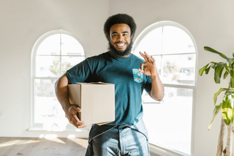 A man dressed in a navy-blue uniform with a black and white patterned headband is inside a room with a textured light blue wall, surrounded by cardboard boxes, some open with packing materials. He is 