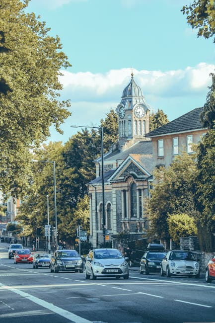 A street scene in Barnet EN5 showing a church with a tall clock tower topped by a spire, surrounded by mature trees and residential buildings. In the foreground, there are multiple parked and moving cars on the road, with some vehicles waiting at a traffic light. To the side of the pavement, a man with a van from Man with Van Barnet is engaged in a home relocation process, lifting a large cardboard box wrapped in plastic and fabric from a trolley or dolly. The area appears well-lit with natural daylight, and the surroundings suggest an active moving and furniture transport environment typical of house removals and packing services. The background includes a clear sky and the church’s architecture, indicating a peaceful suburban or town centre setting. Such logistics are part of comprehensive moving services offered by [COMPANY_NAME], supporting efficient packing, loading, and transportation in residential relocations.